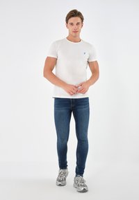 T-shirt bianco a maniche corte con logo blu, abbinato a jeans slim-fit blu scuro e scarpe da ginnastica grigie. Sfondo neutro.