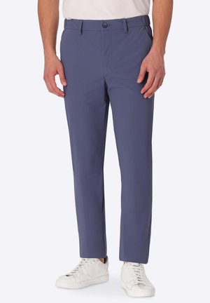 Uomo con pantaloni aderenti blu e scarpe da ginnastica bianche in piedi con le mani rilassate lungo i fianchi davanti a uno sfondo uniforme.