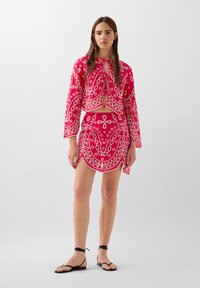 Ensemble deux pièces rose vif composé d'un haut court et d'une jupe assortie, mettant en avant des motifs floraux blancs complexes et des bords festonnés.