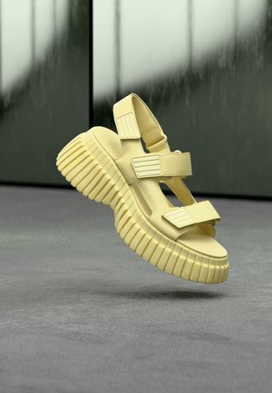 Chunky pale yellow sandal with thick ridged sole and adjustable straps, hovering above a gray surface against a blurred dark background.