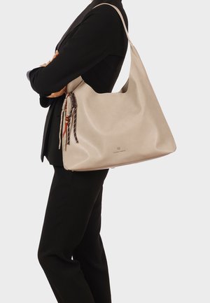 Persona con abito nero che porta una grande borsa a tracolla in pelle beige con nappine decorative su sfondo semplice.