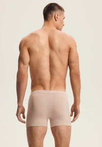 Muscular man with short hair standing, facing away, wearing beige fitted boxer briefs against a plain light background.