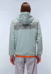 Felpa con cappuccio di colore verde pallido con un motivo grafico strutturato sulla schiena, caratterizzata da un logo e un orlo arancione, abbinata a pantaloni grigi chiari.