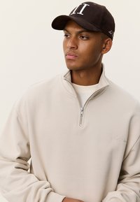 Μπεζ φούτερ με φερμουάρ και ψηλό γιακά, με κεντημένο λογότυπο, σε συνδυασμό με καφέ καπέλο που φέρει λευκές επιγραφές στο μπροστινό μέρος.