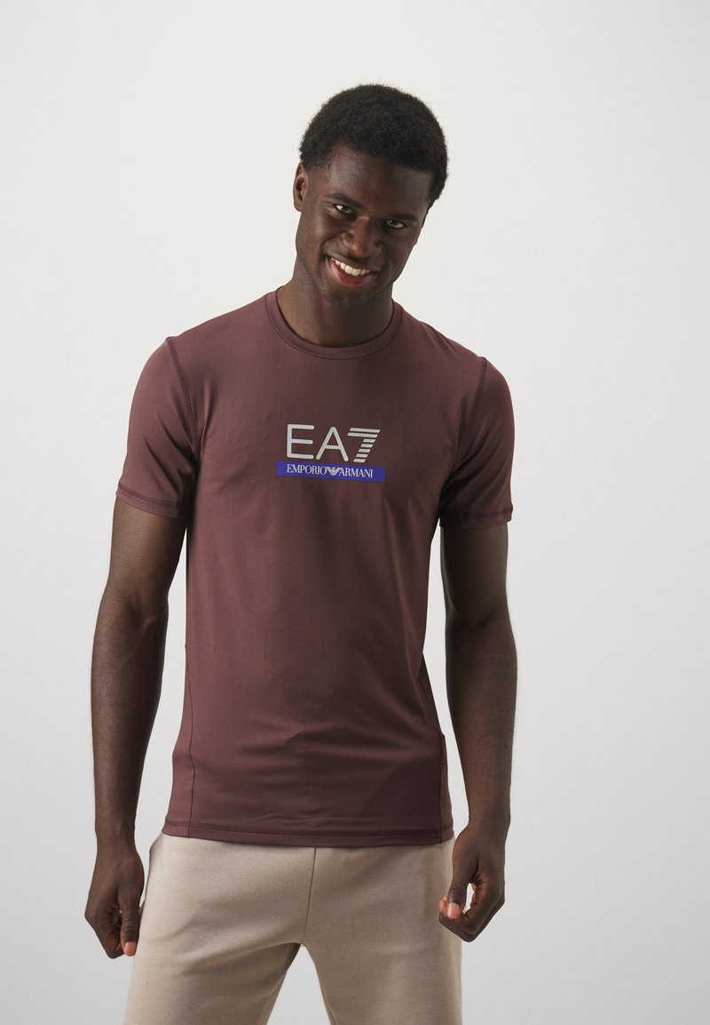 Brun t-skjorte med korte ermer i slim fit, med et sølvfarget EA7-logo og blå aksenttekst. Glatt tekstur og lett materiale.