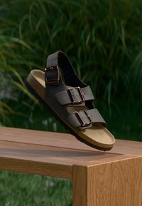 Braune Rutschen-Sandalen mit zwei verstellbaren Riemen, Metall-Schnallen, Korkfußbett und Gummisohle, auf einer Holzoberfläche präsentiert.