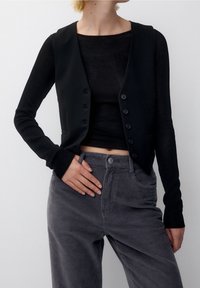 Gilet sans manches noir en tissu lisse avec un devant à boutons, superposé à un haut noir texturé. Associé à un pantalon en velours côtelé gris.