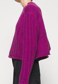 Persona vistiendo un jersey de punto grueso morado con patrones texturizados y pantalones negros, mostrado de perfil contra un fondo liso.