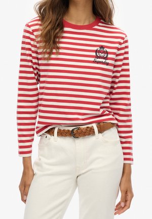 Kobieta ubrana w czerwono-białą bluzkę z długim rękawem w paski z haftowanym logo oraz białe spodnie z brązowym plecionym paskiem.