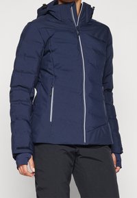 Femme portant une veste matelassée bleu marine avec une capuche, une fermeture éclair à l'avant et des poches latérales zippées, associée à un pantalon noir.