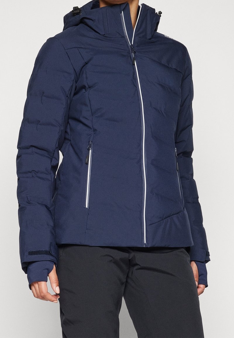 Femme portant une veste matelassée bleu marine avec une capuche, une fermeture éclair à l'avant et des poches latérales zippées, associée à un pantalon noir.