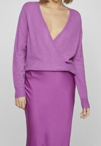 Fialový volný svetr s hlubokým výstřihem do V a žebrovanými manžetami, doplněný elegantní fialovou saténovou sukní s bočními řasením.