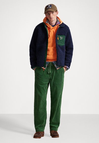 Ung mand iført en brun kasket, orange hoodie, marineblå fleecejakke, ternet skjorte, grønne fløjlspants og brune sko står med hænderne i lommerne.