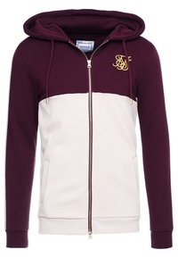 Haut à zip bourgogne et crème avec une grande capuche, des poches kangourou et un accent logo doré sur la poitrine. Tissu doux avec une texture lisse.