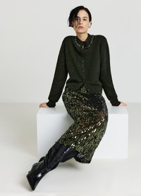 Cardigan vert avec un col orné de perles, associé à une jupe noire à sequins et des bottes noires montantes. Matières texturées avec un design en couches.