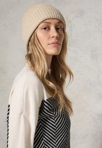 Beanie a coste beige indossato con un maglione a strisce crema e nero. Il maglione presenta un motivo a spina di pesce su un lato. Tessuto liscio.