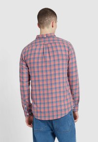 Mężczyzna w długim rękawie, ubrany w czerwono-niebieską koszulę w kratę z zapinanym kołnierzykiem, stojący tyłem, w parze z niebieskimi dżinsami.