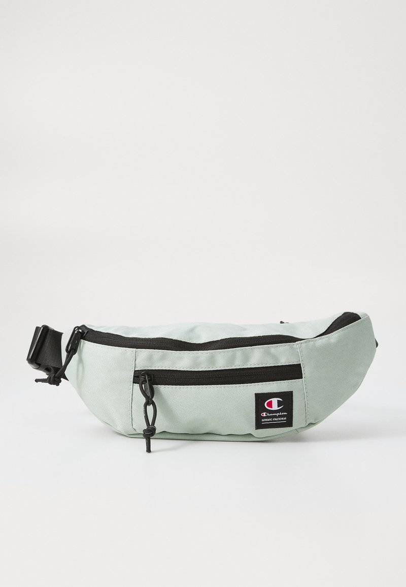 Sac banane en tissu vert avec une forme incurvée, fermeture éclair noire et écusson de logo ; il possède une sangle réglable et une surface texturée.