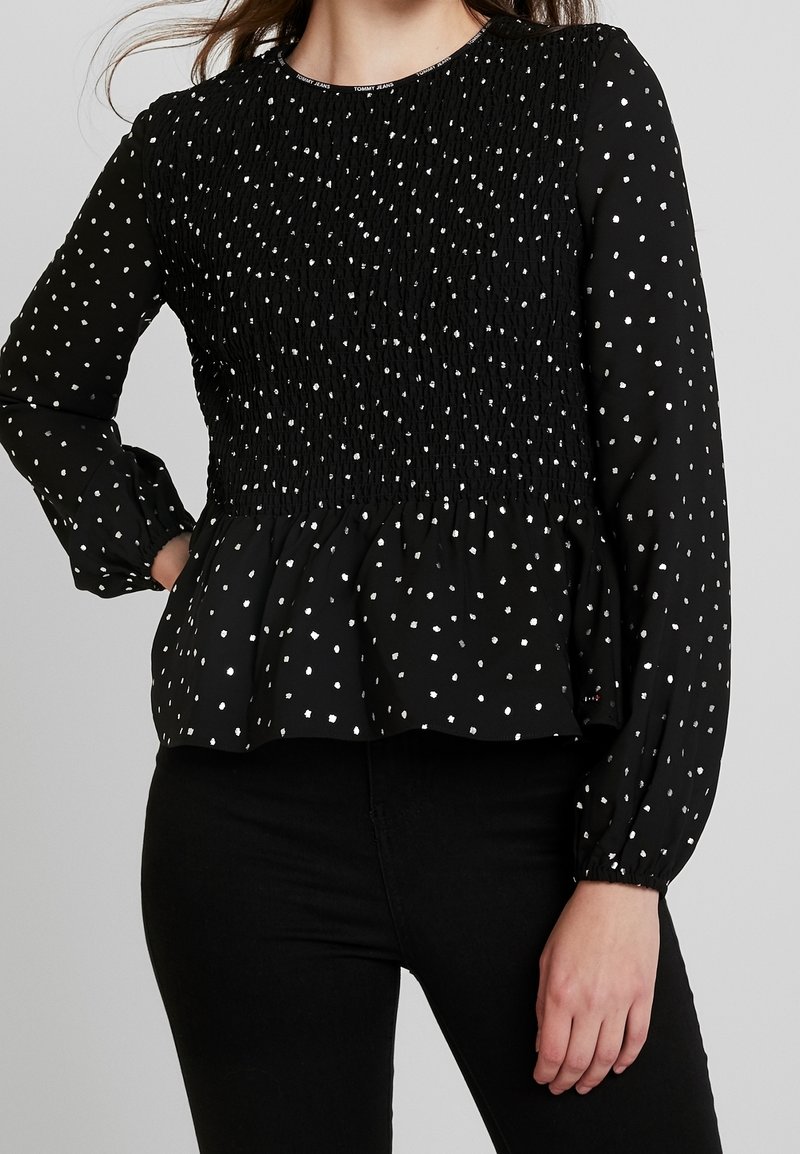 Black blouse with white polka dots, smocked bodice, long puff sleeves, and a flared hem. Made from lightweight fabric with a textured finish.