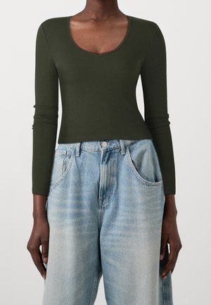 Persoon die een aansluitend, donkergroen, geribd shirt met lange mouwen en een ronde hals draagt, gecombineerd met losse, lichtgewassen blauwe spijkerbroek.