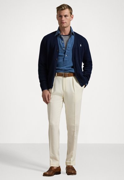 Marinblått stickat cardigan över en ljusblå skjorta med knappar och randig topp, beige byxor, brun bälte och bruna skor.