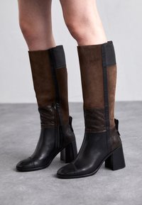 Femme portant des bottes en cuir noires et brunes jusqu'aux genoux avec des talons carrés, se tenant sur un sol gris.