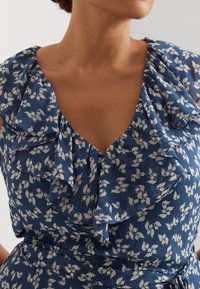 Blue dress with white floral pattern, featuring a deep V-neck and ruffled neckline. Fabric has a textured finish and a fitted waist with tie detail.