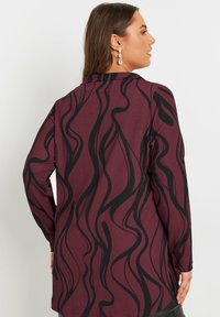 Burgundy långärmad topp med en strukturerad tyg och ett svart vågigt mönster. Designen inkluderar en hög krage och sidokilar.
