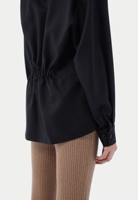 Svart långärmad topp med rynkad detalj vid midjan, kombinerad med ribbade bruna leggings. Slät tyg med en subtil glans.