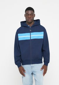 Mørkeblå zip-up hættetrøje med en struktureret lyseblå stribe og hvid detalje, der har en hætte og ribbede manchetter. Båret med lyse jeans.