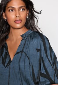 Langærmet, blå bluse lavet af tekstureret stof med en V-udskæring og sorte, abstrakte mønstre. Fremhævet med rynkede detaljer ved kraven.