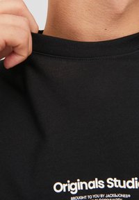 Black cotton t-shirt featuring a round neckline and short sleeves, with white text stating "Originals Studio" on the front.