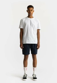 Witte katoenen T-shirt, marineblauwe shorts, witte sokken met rode letters en zwarte sneakers. Model staat tegen een vlakke lichte achtergrond.