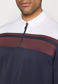 Pánská polo košile s čtvrtinovým zipem. Má bílý límec, pruhy v barvě burgundy a námořnické modři na hrudi a strukturovaný povrch látky.
