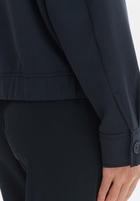 Granatowa bluzka z długim rękawem, o zaokrąglonym dolnym brzegu, z mankietem na guziki i gładką fakturą, zaprojektowana z delikatnymi plisami wzdłuż dołu.