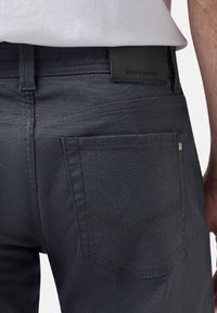 Jeans en denim foncé avec une texture lisse, coupe ajustée et poche arrière avec des coutures discrètes. Étiquette en cuir noir au niveau de la taille.