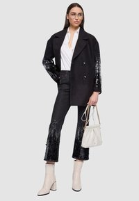 Cappotto nero doppiopetto con maniche decorate con paillettes, abbinato a jeans scuri con sfumatura di paillettes e stivaletti bianchi alla caviglia.