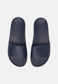 Marina blå slidesandaler tillverkade av texturerad gummi, med en bred rem och en vadderad sula med cirkulära accenter och prickiga mönster.