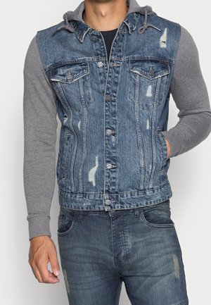 Mand iført blå slidt denimvest med ærmer fra grå hættetrøje og matchende blå slidte jeans, hånd i lommen.