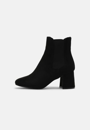 Bottine noire en daim avec talon bloc et panneaux latéraux élastiques, conçue pour un enfilage facile, présentée sur fond blanc.