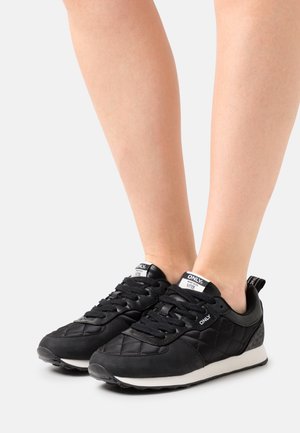 Nohy v černých prošívaných teniskách s bílými podešvemi, černými tkaničkami a bílými značkovými štítky na jazyku a straně, stojící na bílém povrchu.