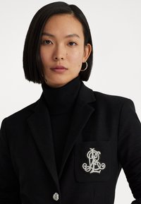 Zwarte blazer met een textuurafwerking, voorzien van een zilver geborduurd logo op de zak en een metallic knoopdetail. Draagt over een zwarte coltrui.