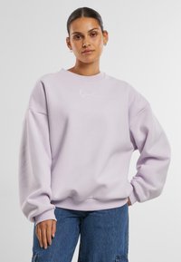 Lavenders spalvos per didelis džemperis, pagamintas iš minkštos medžiagos, su apvalia iškirpte, nusileidusiomis petnešomis ir balta išsiuvinėta logotipu.