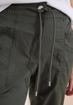 Tmavě zelené kalhoty s šňůrkou a viditelným šitím, nošené s zastrčenou bílou košilí, zobrazující přední kapsy a knoflík v pase.