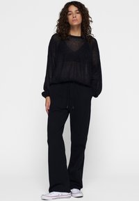 Schwarzer, transparenter Pullover mit langen Ärmeln und lockerer Passform; kombiniert mit locker sitzenden schwarzen Hosen und weißen Sneakers mit roten Akzenten.