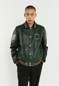 Veste varsity en cuir vert avec des manches en cuir noir, col côtelé à rayures marron et noir, présentant des détails de badge et des boutons-pression.