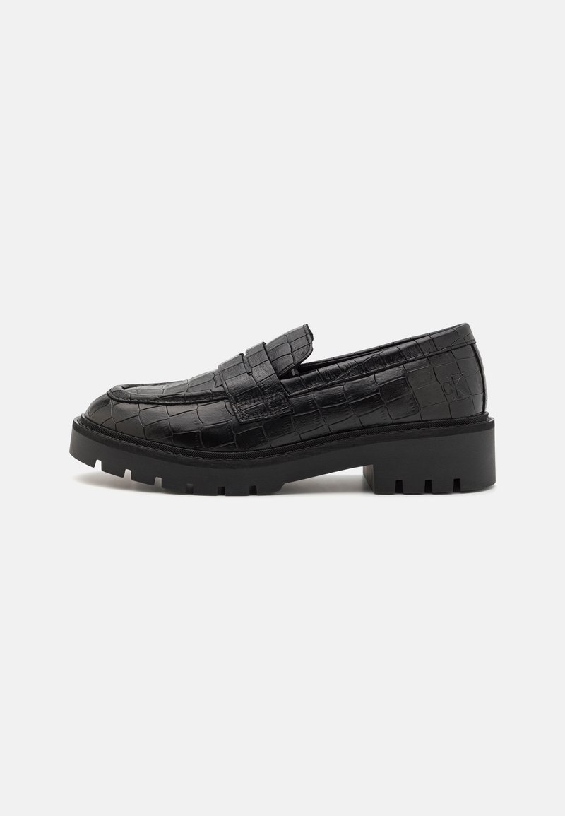 Mocassin en cuir noir à motif crocodile, semelle épaisse et talon bas, présenté de profil sur un fond neutre.