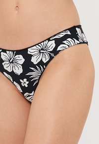 Melna peldbikini apakšda ar baltu ziedu rakstu, ieskaitot hibiskus un palmas lapas, elastīgu jostu un gludu auduma tekstūru.