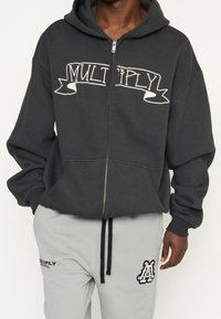 Sweat à capuche gris à fermeture éclair avec une poche avant, portant le texte blanc "MULTIPLY". Associé à un pantalon de survêtement gris clair affichant un logo.