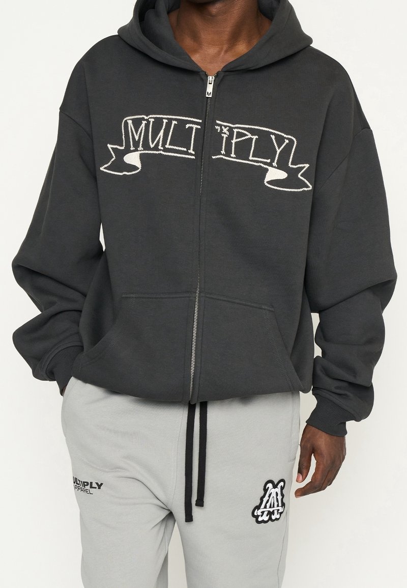 Sweat à capuche gris à fermeture éclair avec une poche avant, portant le texte blanc "MULTIPLY". Associé à un pantalon de survêtement gris clair affichant un logo.